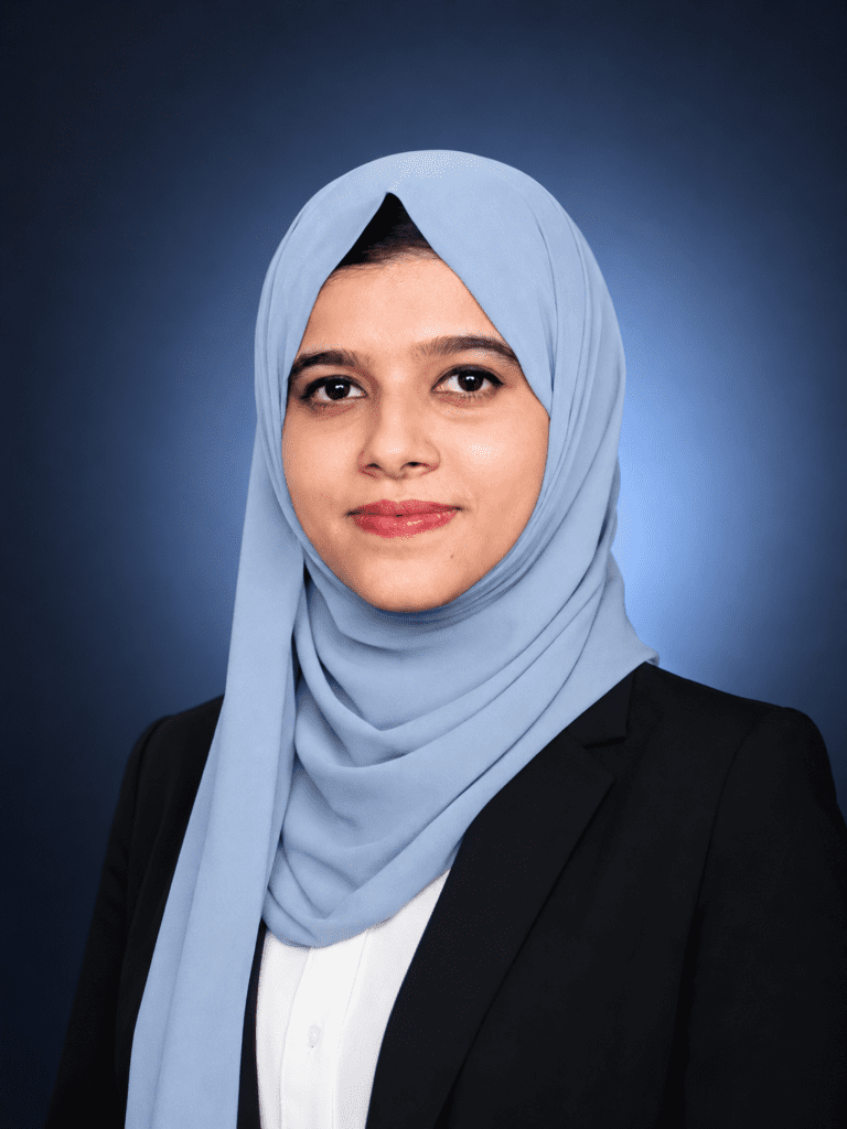 Professional portrait of a young Muslim woman in a light blue hijab and black blazer, smiling confidently against a navy blue studio background. Ideal branding image for a digital marketing consultant in Kerala, showcasing professionalism, confidence, and a modern corporate style.